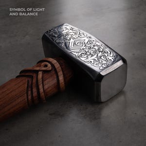 May include: A detailed close-up of a hammer with a polished metal head and a carved wooden handle. The metal head features intricate floral and geometric engravings. The text "SYMBOL OF LIGHT AND BALANCE" is at the top left.