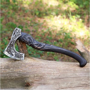 May include: A hand-forged axe with a black wooden handle and a silver blade. The blade is decorated with an intricate design of a dragon and other mythical creatures. The axe is resting on a wooden log.