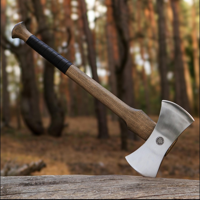 K&ouml;nnte beinhalten: Eine Doppelaxt mit Holzgriff und poliertem Stahlkopf. Der Griff ist mit schwarzem Leder umwickelt. Die Axt liegt auf einem Baumstamm in einer Waldumgebung.