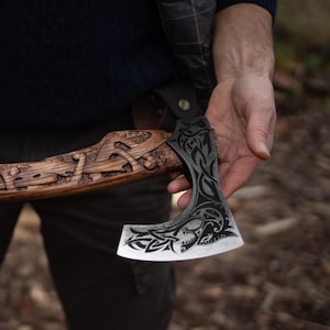 May include: Axe with a wooden handle and a metal blade. The handle is carved with intricate designs, and the blade features a dark, etched pattern. The axe is held in a person's hand, showcasing the craftsmanship and detail.