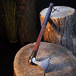 May include: A handcrafted axe with a dark wooden handle and a silver-colored blade. The handle is wrapped in black leather near the head and the top. The axe is resting on a wooden surface.