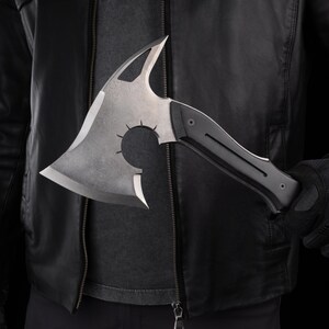 Puede incluir: Un hacha tomahawk plateada y negra. La cabeza del hacha presenta un recorte circular con l&iacute;neas radiales y una hoja curva. El mango negro es de material compuesto. El hacha se sostiene frente a una chaqueta de cuero negra.