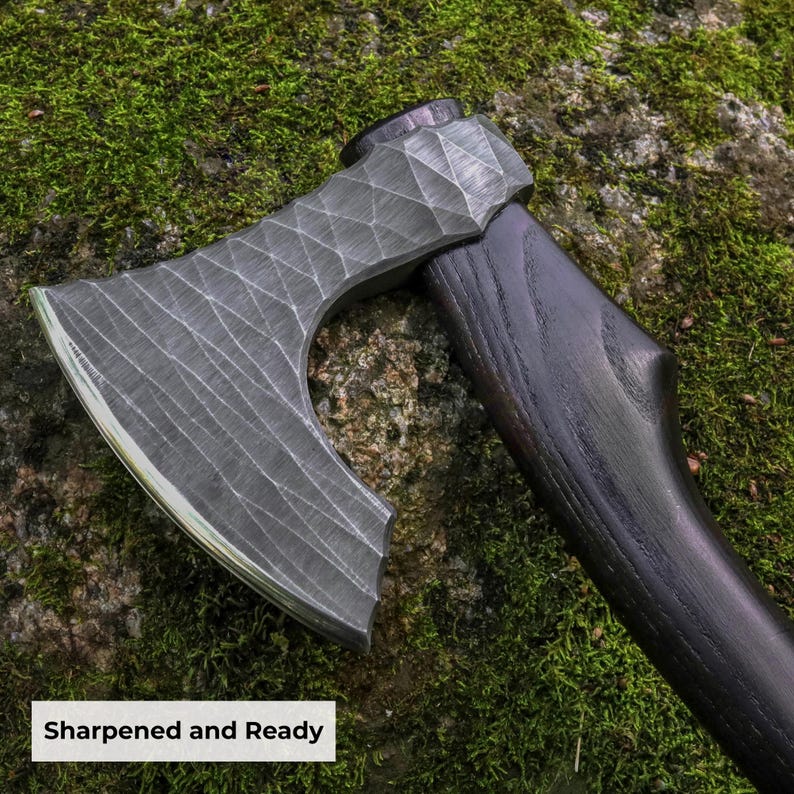 May include: A hand axe featuring a dark, textured metal head with a distinct cross-hatched, faceted pattern and a sharp, curved edge. The handle is crafted from dark, stained wood with a visible grain. The text Sharpened and Ready is visible.