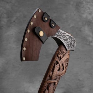 Pu&ograve; includere: Un'ascia artigianale con un manico in legno intagliato. La testa dell'ascia &egrave; in metallo con un motivo decorativo ed &egrave; ricoperta da un fodero in pelle marrone fissato con rivetti e cinghie. Il manico &egrave; di colore marrone scuro.