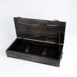 May include: A rectangular wooden box with a dark, distressed wood grain finish and an open hinged lid. The interior is lined with black fabric and features two black leather straps with silver-toned metal buckles for securing items inside.