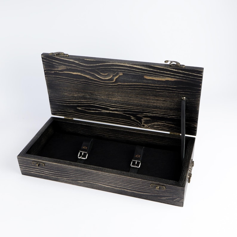 May include: A rectangular wooden box with a dark, distressed wood grain finish and an open hinged lid. The interior is lined with black fabric and features two black leather straps with silver-toned metal buckles for securing items inside.