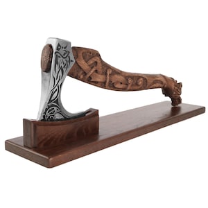 May include: A hand-carved wooden axe with a detailed design of a dragon and intricate knotwork. The axe head is made of metal and has a sharp edge. The axe is mounted on a wooden stand.