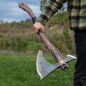 Puede incluir: Un hacha decorativa detallada con un mango de madera y una hoja de metal. El mango presenta intrincadas tallas y está envuelto con cinta adhesiva negra. La hoja tiene un diseño azul y dorado. El hacha se sostiene frente a un fondo de hierba verde.
