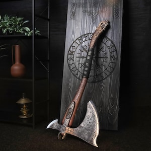May include: A decorative battle axe with a silver-toned metal head featuring intricate etched patterns and a wooden handle wrapped in black leather. The handle is topped with a carved metal pommel and rests against a dark wooden plaque with runes.