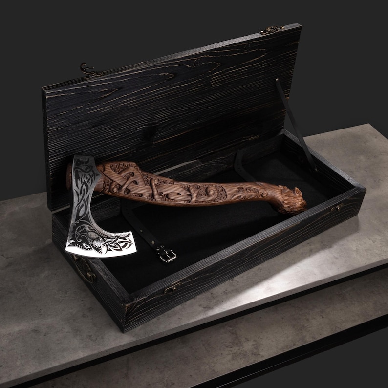 Pu&ograve; includere: Un'ascia decorativa con un manico in legno intagliato e una lama argentata, presentata in una scatola di legno nera. Il manico presenta intagli intricati e la lama ha un design dettagliato. La scatola &egrave; foderata con tessuto nero.