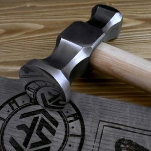 K&ouml;nnte beinhalten: Ein Stahlhammer mit Holzgriff. Der Hammerkopf ist dunkelgrau, der Griff hellbraun. Der Hammer liegt auf einer grauen Holzoberfl&auml;che mit ge&auml;tzten Designs. Das Ganze liegt auf einer Holzoberfl&auml;che.