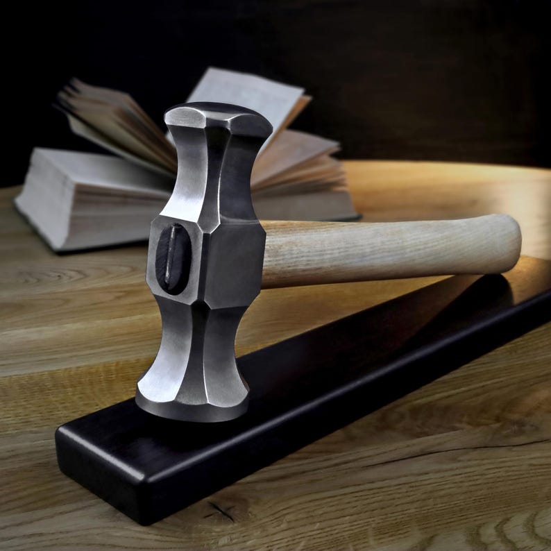 K&ouml;nnte beinhalten: Ein Stahlhammer mit hellem Holzgriff liegt auf einer schwarzen Oberfl&auml;che. Der Hammerkopf hat ein einzigartiges, facettiertes Design. Im Hintergrund liegt ein offenes Buch auf einer Holzoberfl&auml;che.