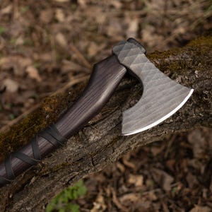 Könnte beinhalten: Eine Handaxt mit einem dunkelbraunen Holzgriff, der mit einem schwarzen Lederband umwickelt ist. Der Metall-Axtkopf hat eine texturierte, facettierte Oberfläche mit einer polierten, scharfen silbernen Schneide. Das Design umfasst eine gebogene Klinge.