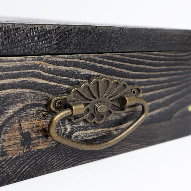 May include: A rectangular wooden box featuring a dark, charred wood grain finish and a decorative antique brass-toned metal drawer pull. The handle is attached with two small screws and has a scalloped, fan-like backplate design.