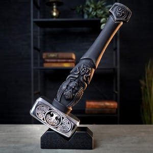 Puede incluir: Un martillo gris oscuro detallado con intrincadas tallas de caras y diseños de nudos celtas. El cabezal del martillo es plateado con un diseño de árbol de la vida. El mango es negro y el martillo se exhibe sobre un soporte negro.