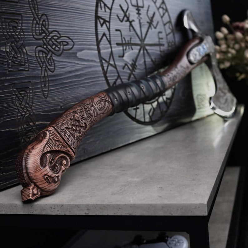 May include: A handcrafted axe with a wooden handle featuring intricate carvings and a leather-wrapped grip. The axe head is made of metal. The axe is displayed against a wooden backdrop with etched symbols.
