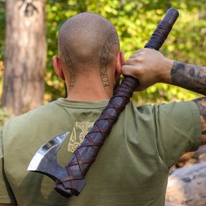 May include: A Viking-style axe with a dark metal blade and a wooden handle wrapped in brown leather. The handle has a cross-stitched leather design. The axe is held over the shoulder.