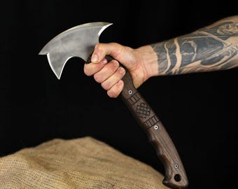 May include: A handcrafted axe with a curved, silver blade and a dark brown wooden handle. The handle has decorative carvings and metal rivets. The axe is held against a black background.