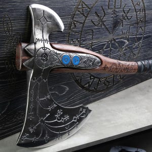 May include: A detailed, silver-colored axe with intricate engravings and a wooden handle. The handle features blue accents and decorative metalwork. The axe is mounted on a dark wooden surface with carved symbols, showcasing a blend of craftsmanship and design.