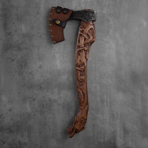 Pu&ograve; includere: Un'ascia artigianale con una testa in metallo scuro e un manico in legno intagliato. Il manico presenta intagli intricati e una testa di lupo alla base. Un fodero in pelle marrone protegge la testa dell'ascia. L'ascia &egrave; un oggetto decorativo.