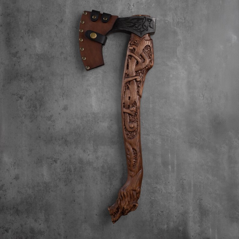 Pu&ograve; includere: Un'ascia artigianale con una testa in metallo scuro e un manico in legno intagliato. Il manico presenta intagli intricati e una testa di lupo alla base. Un fodero in pelle marrone protegge la testa dell'ascia. L'ascia &egrave; un oggetto decorativo.