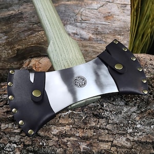 K&ouml;nnte beinhalten: Axt mit einer silbernen Klinge und einem gr&uuml;nen Holzgriff. Die Klinge hat ein dekoratives kreisf&ouml;rmiges Emblem und wird durch eine schwarze Lederscheide gesch&uuml;tzt, die mit Messingnieten und einem Riemen befestigt ist.