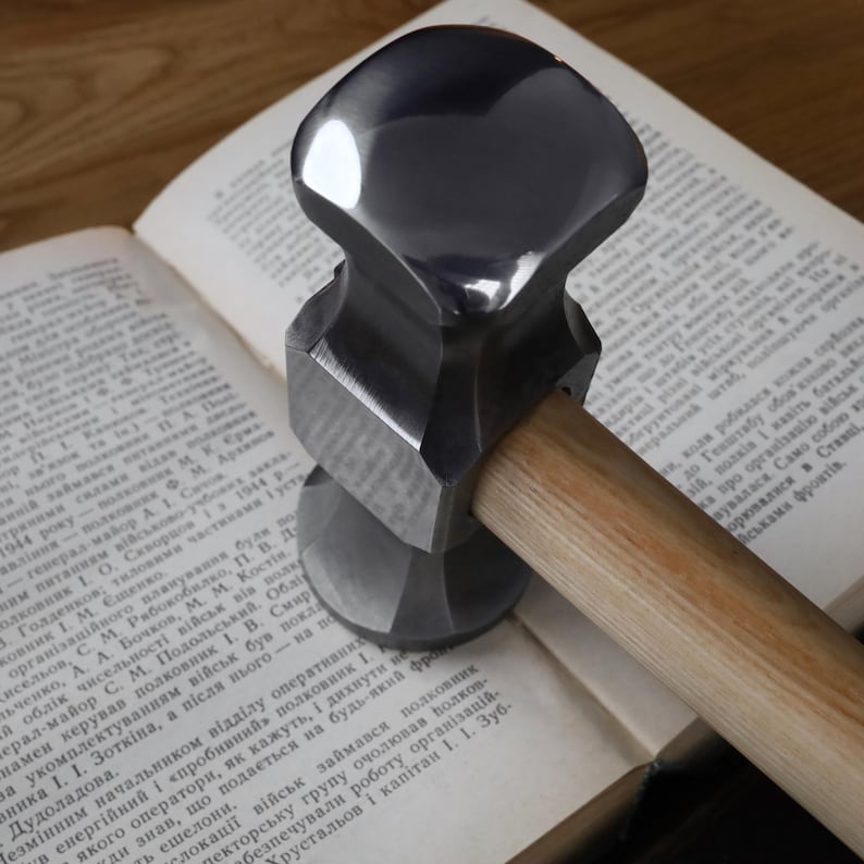 K&ouml;nnte beinhalten: Ein polierter Stahlhammer mit Holzgriff liegt auf einem offenen Buch. Der Hammerkopf hat eine abgerundete Oberseite und einen facettierten unteren Abschnitt. Der Griff ist aus hellem Holz.