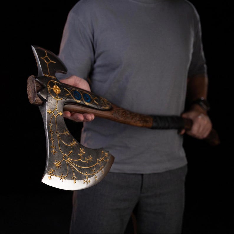 May include: A double-bit axe featuring a dark, weathered metal head adorned with intricate gold-colored etched patterns and two circular blue inlays. The handle is crafted from dark wood with a textured grip section near the axe head.