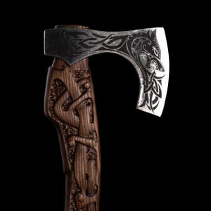 Pu&ograve; includere: Ascia con manico in legno scuro con figure scolpite e una lama in acciaio. La lama presenta intricati disegni incisi. L'ascia &egrave; posta su uno sfondo nero.