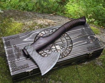 May include: A dark-handled axe with a textured blade rests on a wooden box. The box is dark gray with Celtic knot designs and a circular rune pattern. The axe handle is wrapped in brown leather.