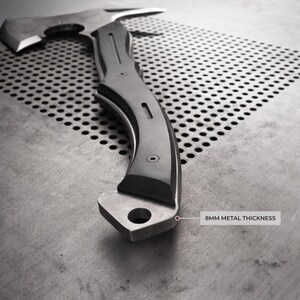 Puede incluir: Un hacha plateada con un mango curvo y una hoja afilada. El mango tiene un agarre oscuro y texturizado. El texto "8MM METAL THICKNESS" es visible. El hacha est&aacute; dise&ntilde;ada para uso intensivo.