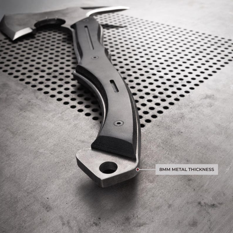 Puede incluir: Un hacha plateada con un mango curvo y una hoja afilada. El mango tiene un agarre oscuro y texturizado. El texto "8MM METAL THICKNESS" es visible. El hacha est&aacute; dise&ntilde;ada para uso intensivo.