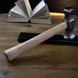 K&ouml;nnte beinhalten: Ein Metallhammer mit hellem Holzgriff liegt auf einem dunklen Holzsockel. Der Hammerkopf ist dunkelgrau und der Griff hellbraun. Im Hintergrund ist ein offenes Buch zu sehen.