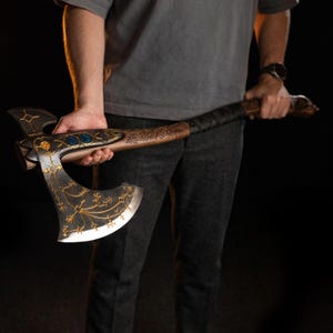 May include: A double-bitted axe with a dark wooden handle and a metal head featuring intricate gold-colored filigree patterns, blue circular inlays, and a sharp, polished edge. The handle is wrapped in black material and displays carved markings.
