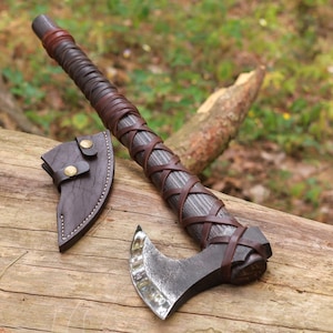 May include: A handcrafted axe with a dark wooden handle wrapped in brown leather. The axe head is made of metal and has a curved blade. A brown leather sheath with brass buttons is included. The axe is displayed on a wooden surface.