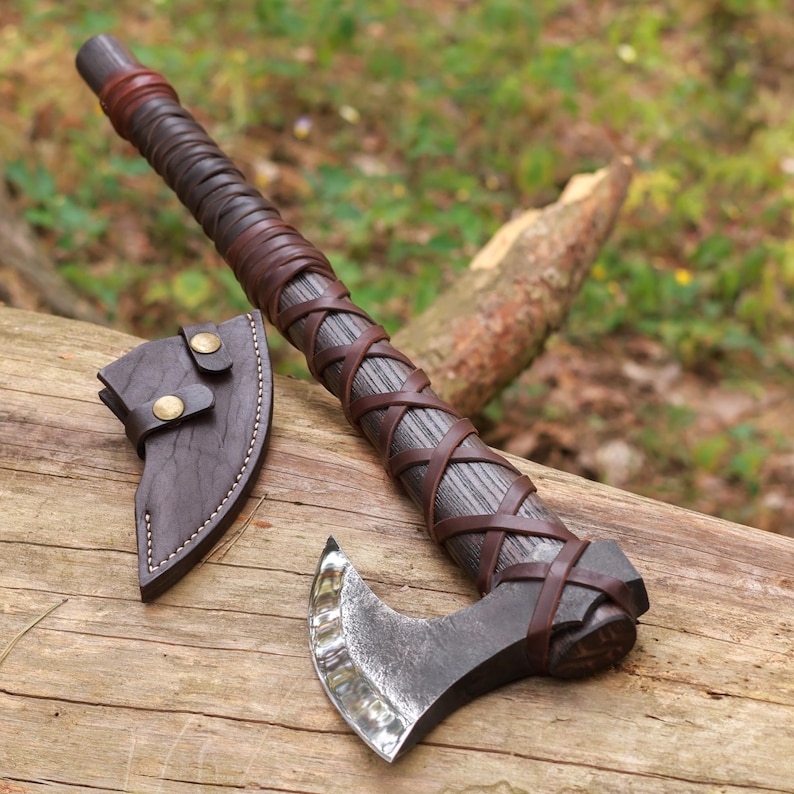 May include: A handcrafted axe with a dark wooden handle wrapped in brown leather. The axe head is made of metal and has a curved blade. A brown leather sheath with brass buttons is included. The axe is displayed on a wooden surface.