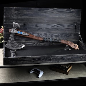 May include: A detailed, decorative axe with a silver blade and a wooden handle, displayed in a black wooden box. The axe features intricate carvings and blue accents. The handle is wrapped in leather. The box is open, revealing the axe's full length.