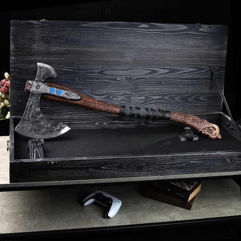 May include: A detailed, decorative axe with a silver blade and a wooden handle, displayed in a black wooden box. The axe features intricate carvings and blue accents. The handle is wrapped in leather. The box is open, revealing the axe's full length.