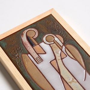 May include: A framed ceramic tile with a stylized design of two figures in shades of brown, white, and green. The tile is set against a green background with a brown frame.
