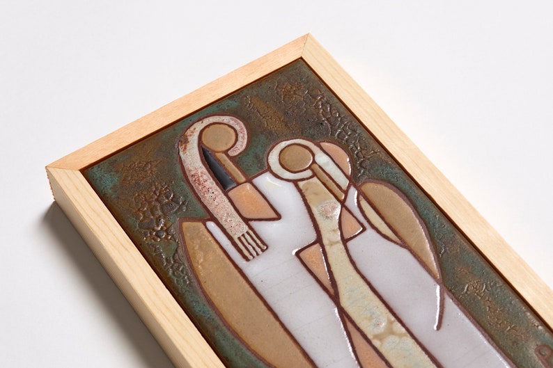 May include: A framed ceramic tile with a stylized design of two figures in shades of brown, white, and green. The tile is set against a green background with a brown frame.