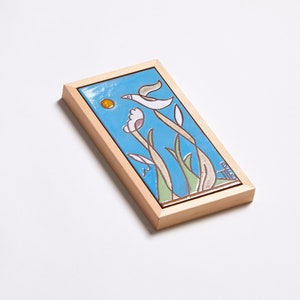 Puede incluir: Un azulejo de cer&aacute;mica enmarcado con un fondo azul, un p&aacute;jaro blanco volando y flores amarillas y rosas. El azulejo est&aacute; enmarcado en un marco de madera claro.
