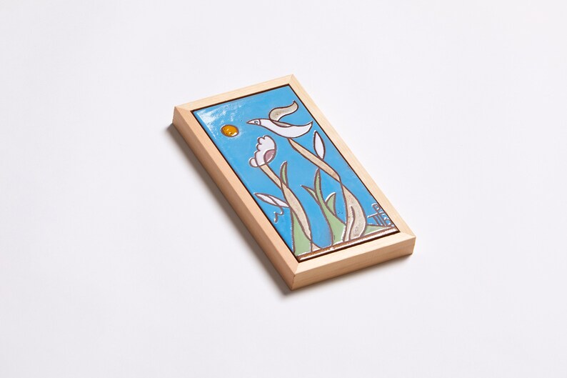 Puede incluir: Un azulejo de cer&aacute;mica enmarcado con un fondo azul, un p&aacute;jaro blanco volando y flores amarillas y rosas. El azulejo est&aacute; enmarcado en un marco de madera claro.