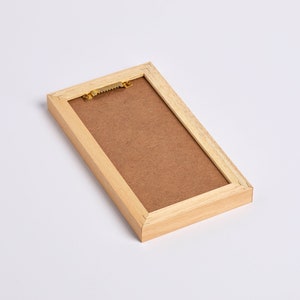 Peut inclure: Un cadre photo en bois avec un fond en carton brun et un crochet de suspension en laiton. Le cadre est brun clair et a un design simple.