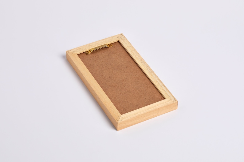 Peut inclure: Un cadre photo en bois avec un fond en carton brun et un crochet de suspension en laiton. Le cadre est brun clair et a un design simple.