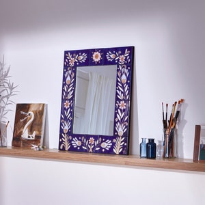 Puede incluir: Un espejo decorativo con un marco azul que presenta un diseño floral en blanco y rosa. El espejo está colgado en un estante con una planta en maceta, una obra de arte y pinceles.