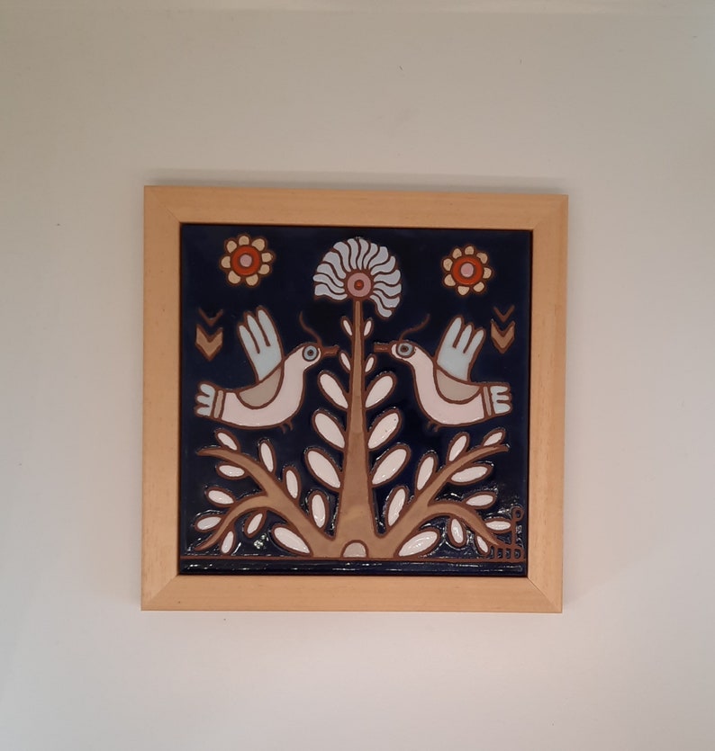 May include: A framed ceramic tile with a blue background featuring two white birds perched on a stylized tree with white leaves. The tree has three red and orange flowers. The tile is framed in a light wood frame.