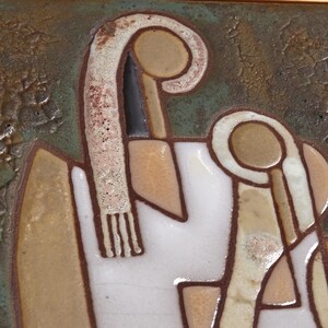 May include: A ceramic tile with a stylized design of two figures. The figures are outlined in brown and have a white background. The tile has a textured, brown background.