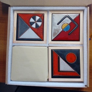 K&ouml;nnte beinhalten: Vier Keramikfliesen mit geometrischen Mustern in einer Holzkiste. Die Fliesen sind in Schwarz, Wei&szlig;, Rot und Blau gehalten. Die Designs beinhalten einen Kreis mit einem kleineren Kreis im Inneren, eine geschwungene Linie, ein Dreieck und einen Kreis.