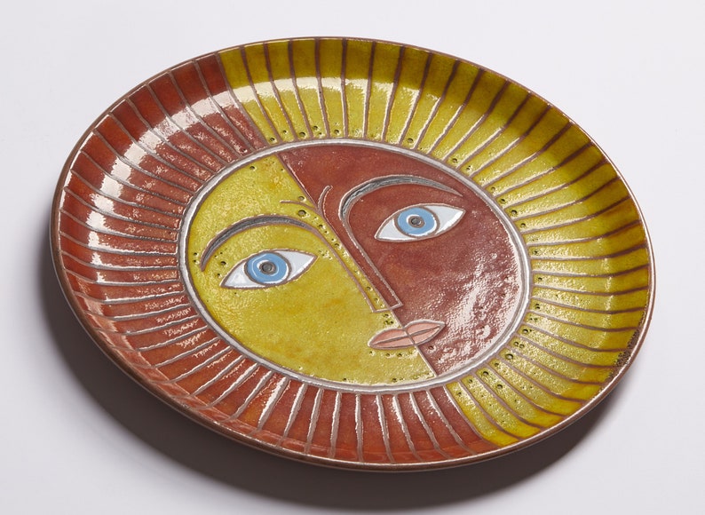 May include: A ceramic plate with a stylized sun face design. The sun has a yellow and red face with blue eyes and a pink mouth. The plate has a red and yellow rim with a sunburst design.
