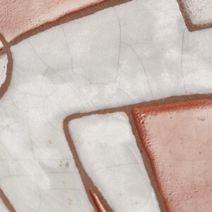 Op de afbeelding: Close-up van een keramische tegel met een witte glazuur en bruine lijnen die een geometrisch patroon vormen.